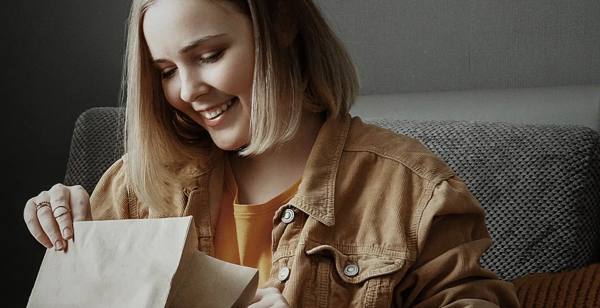 A woman on a couch holds a brown paper bag, opening it to reveal a surprise gift inside