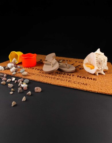 An image of a TerraGuard Doormat—a customizable welcome doormat with an agent's name, phone number, and website printed on it. The doormat features a pair of beige flip-flops, two small beach buckets (one yellow, one orange) filled with pebbles, a large seashell, and scattered pebbles, set against a black background.